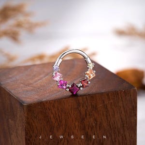 May include: A silver-colored nose ring with a row of colorful square-cut gemstones. The gemstones are arranged in a circular pattern, showcasing shades of pink, purple, orange, and red. The word "JEWSEEN" is printed at the bottom of the image.
