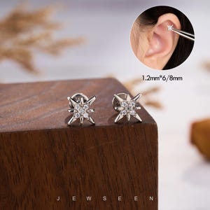 May include: A pair of silver star-shaped stud earrings with small, clear gemstones. The earrings are displayed on a wooden surface. The text "JEWSEEN" is at the bottom of the image. The earrings measure 1.2mm x 6/8mm.