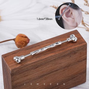 May include: A silver industrial barbell earring with a bone-shaped design. The earring measures 1.6mm x 38mm and is displayed on a wooden block. The word "JEWSEEN" is printed at the bottom of the block.