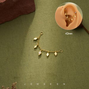 May include: A gold-colored ear chain with dangling diamond-shaped charms. The chain is 42mm long and features a small ring at each end for attachment. The design includes a small, clear gem in one of the charms. The word "JEWSEEN" is at the bottom.