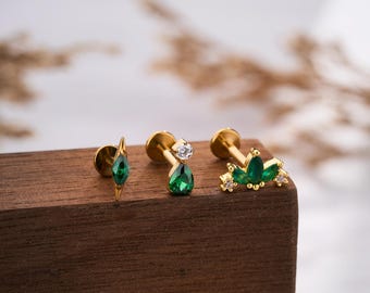 May include: Three gold-colored earrings with green and clear gemstones are displayed on a wooden block. The earrings feature various designs, including a teardrop, a diamond shape, and a cluster of gems. The word "JEWSEEN" is printed at the bottom.