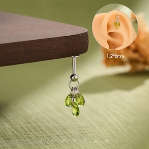 May include: A silver-colored, dangling body piercing with a cluster of green, teardrop-shaped gems. The piercing is suspended from a small ball and ring. The image also shows a close-up of the piercing in an ear, with the text "1.2*6mm".