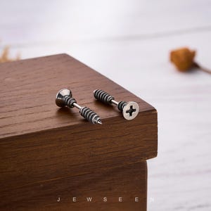 May include: Two silver-toned screw-shaped stud earrings with black detailing. One earring has a flat head with a cross-shaped indentation, and the other has a pointed end. The earrings are displayed on a dark wooden surface.