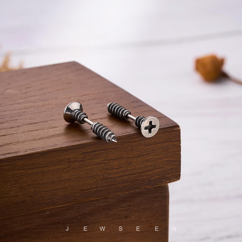 May include: Two silver-toned screw-shaped stud earrings with black detailing. One earring has a flat head with a cross-shaped indentation, and the other has a pointed end. The earrings are displayed on a dark wooden surface.