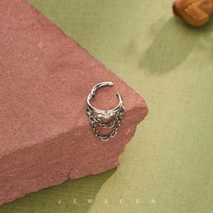 May include: Silver nose ring featuring a star-shaped centre and hanging chains. The jewellery is presented on a red-brown surface against a green backdrop. The word "JEWSEEN" is printed at the bottom of the image.