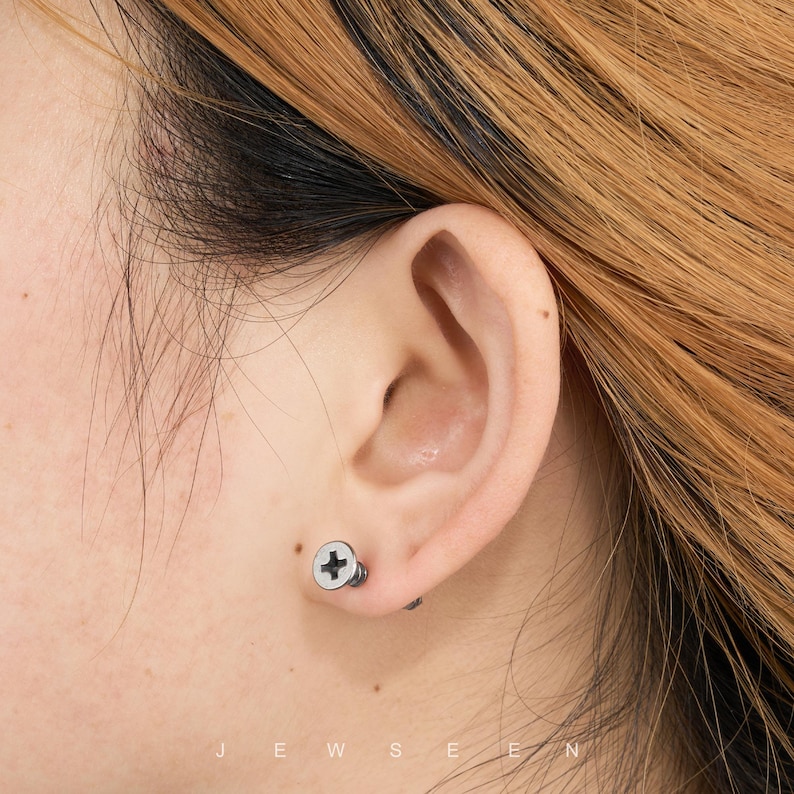 May include: A close-up of an ear with a silver screw-shaped earring. The earring has a white head with a black cross design. The word "JEWSEN" is printed at the bottom of the image.