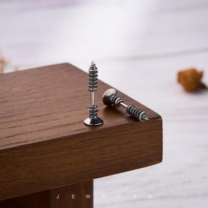 May include: Two silver-toned screw-shaped stud earrings. One earring stands upright, while the other lies on its side. The earrings are on a dark brown wooden surface. The word "JEWSEEN" is at the bottom of the image.