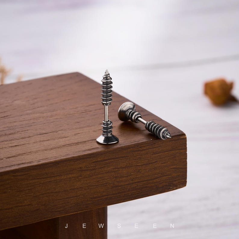 May include: Two silver-toned screw-shaped stud earrings. One earring stands upright, while the other lies on its side. The earrings are on a dark brown wooden surface. The word "JEWSEEN" is at the bottom of the image.