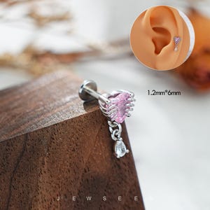 May include: A silver body piercing with a pink heart-shaped gem and a dangling clear gem. The piercing is 1.2mm by 6mm.