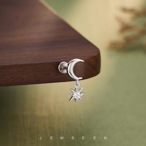 Puede incluir: Pendiente de plata con un diseño de media luna y un colgante de estrella. La estrella tiene una gema transparente en el centro. El pendiente se muestra sobre una superficie de madera oscura y un fondo verde. La palabra "JEWSEEN" está en la parte inferior.