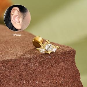 May include: A gold-colored stud earring with a square-cut, clear gemstone at the center, flanked by two smaller, marquise-cut gems. The earring is shown on a textured, reddish-brown surface. The earring post is 1.2mm by 8mm.