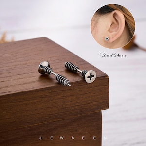 May include: Two silver-toned screw-shaped earrings with black detailing. The earrings are displayed on a wooden surface. The text "JEW SEE" is at the bottom of the image. The earrings measure 1.2mm x 24mm.