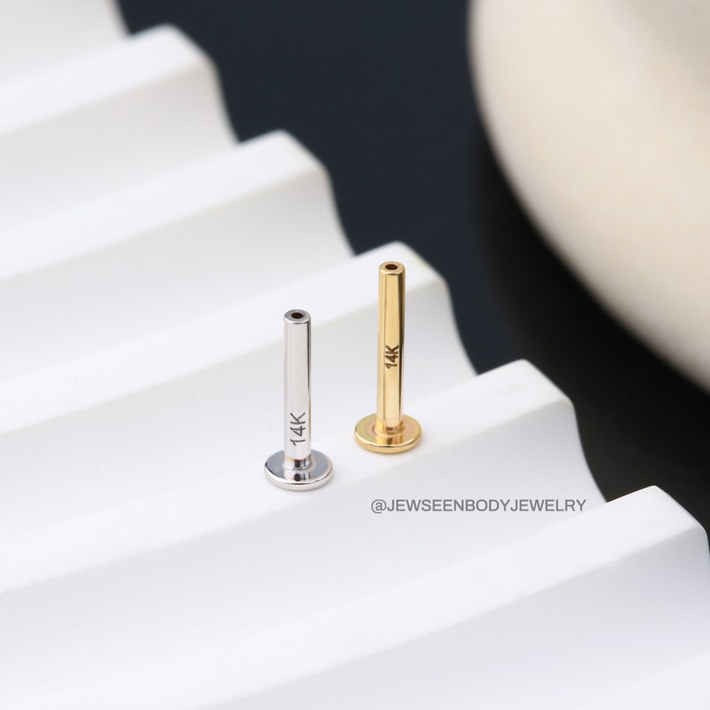 Op de afbeelding: Twee 14 karaats gouden lichaamsieraden, een zilveren en een gouden, op een wit oppervlak. De sieraden hebben de vorm van een kleine, dunne staaf met een platte, ronde basis.