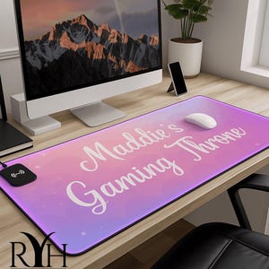 May include: A rectangular gaming mouse pad with a gradient pink and purple background. The words "Maddie's Gaming Throne" are written in white script. A wireless charging pad and a computer mouse are on the pad.