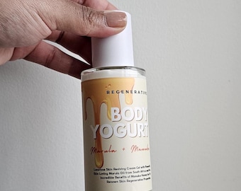 Op de afbeelding: Een heldere glazen fles body yoghurt met een witte dop. Het etiket bevat de woorden "BODY YOGURT" en "Marula + Manuka" in goud. De fles wordt door een persoon vastgehouden.