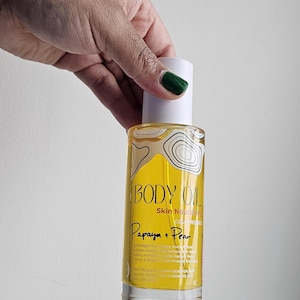 May include: A clear glass bottle of body oil with a white cap, held by a hand with green nail polish. The label reads "BODY OIL" and "Papaya + Pear". The oil appears yellow.