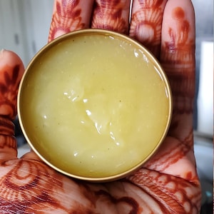 May include: A small, round, gold-colored container filled with a light yellow, semi-solid substance. The container is held in a hand decorated with intricate henna designs in shades of brown.