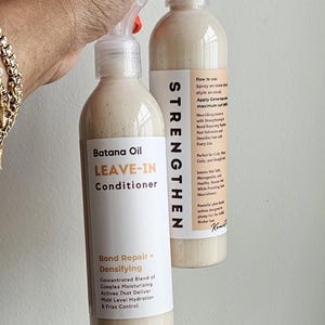 May include: Two clear plastic spray bottles containing beige Batana Oil Leave-In Conditioner. The labels feature black and orange text including the words STRENGTHEN, Bond Repair + Densifying, and instructions for use on curly, wavy, coily, and straight hair.
