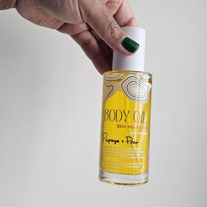 May include: A bottle of Body Oil, labeled "Papaya + Pear," held by a person. The bottle has a white cap and a yellow label with text and a graphic. The oil is skin nourishing and cold pressed.