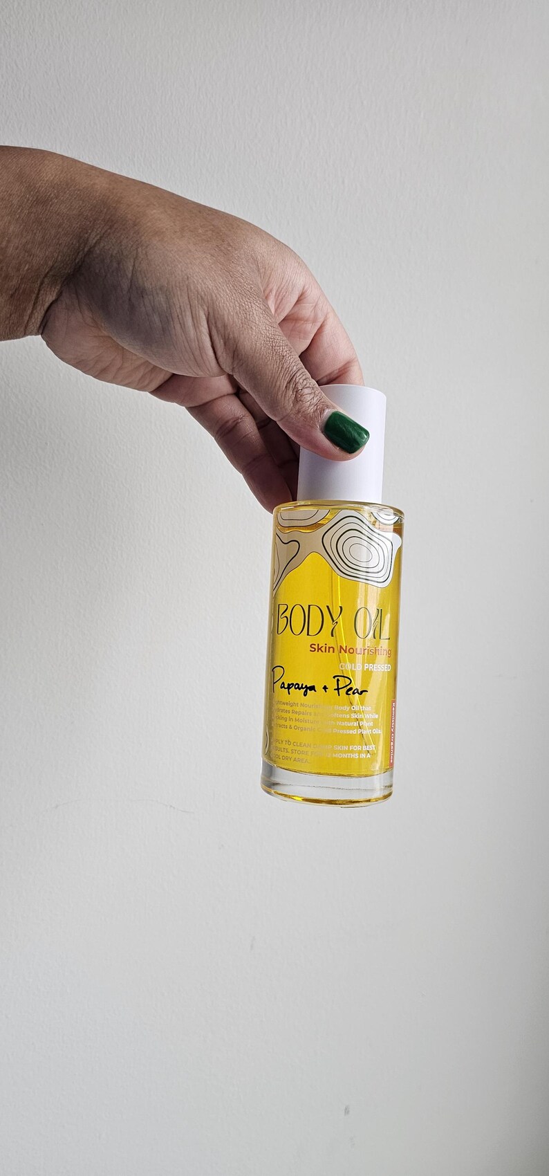 May include: A bottle of Body Oil, labeled "Papaya + Pear," held by a person. The bottle has a white cap and a yellow label with text and a graphic. The oil is skin nourishing and cold pressed.