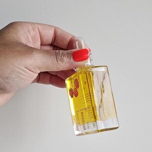 Puede incluir: Una botella de vidrio transparente y rectangular llena de l&iacute;quido amarillo, sostenida por una mano. La botella tiene una tapa cuentagotas y una etiqueta con texto y una ilustraci&oacute;n de frambuesa. La botella mide aproximadamente 7.6 cm de alto.