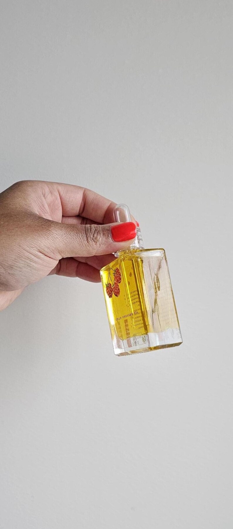 Puede incluir: Una botella de vidrio transparente y rectangular llena de l&iacute;quido amarillo, sostenida por una mano. La botella tiene una tapa cuentagotas y una etiqueta con texto y una ilustraci&oacute;n de frambuesa. La botella mide aproximadamente 7.6 cm de alto.