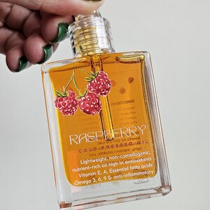 Puede incluir: Una botella cuadrada transparente de aceite de frambuesa con tapa dorada. La etiqueta presenta la palabra "RASPBERRY" y un gr&aacute;fico de tres frambuesas rojas. La botella contiene un l&iacute;quido dorado.