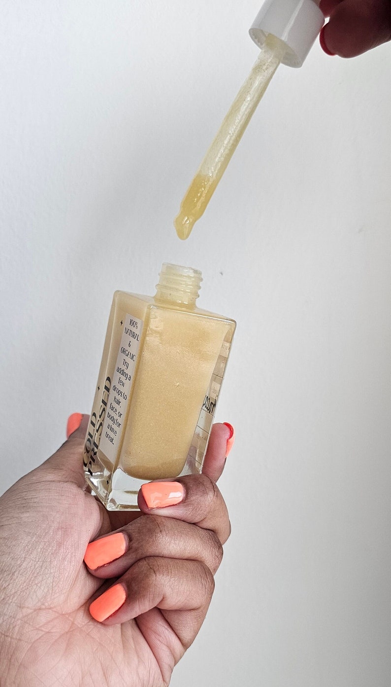 May include: A clear glass bottle with a gold liquid inside. The bottle has a black label with the text 'GOLD BLESSED' and '100% NATURAL & ORGANIC. Try adding a few drops to face, hair, or body for a luxe treat.'