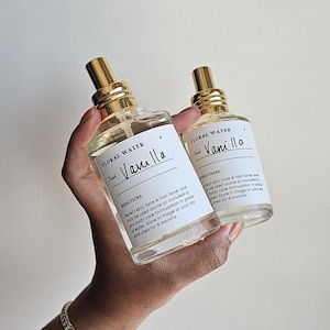 May include: Two clear glass bottles of floral water with gold-coloured spray tops are held in a hand. Each bottle has a white label with the word "Vanilla" and the text "Floral Water". The bottles are for skin, face, and hair.