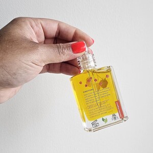 Puede incluir: Una botella rectangular transparente llena de l&iacute;quido amarillo, sostenida por una mano con esmalte de u&ntilde;as rojo. La botella tiene una tapa transparente y una etiqueta con texto e ilustraciones de bayas. La etiqueta dice "Kentry Organics".