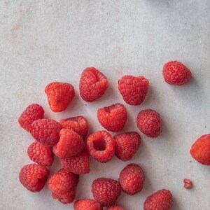 Puede incluir: Un conjunto de frambuesas rojas frescas y maduras esparcidas sobre una superficie clara. Las bayas tienen varios tama&ntilde;os y formas, mostrando sus superficies texturizadas y su color vibrante. La imagen es una vista superior, destacando la belleza natural de la fruta.