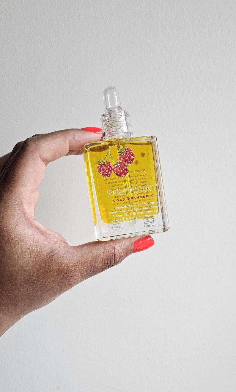 Puede incluir: Una botella cuadrada de vidrio transparente llena de l&iacute;quido amarillo, etiquetada como "RASPBERRY COLD PRESSED OIL". La botella presenta un cuentagotas transparente y un gr&aacute;fico de frambuesas rojas. El texto destaca las propiedades antioxidantes y antiinflamatorias del aceite.