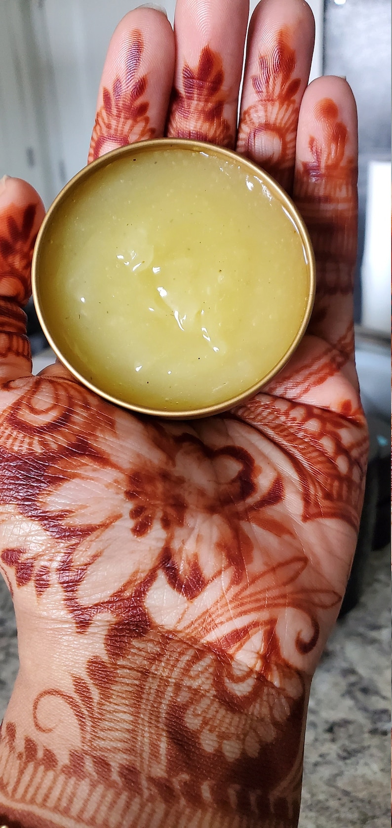 May include: A small, round, gold-colored container filled with a light yellow, semi-solid substance. The container is held in a hand decorated with intricate henna designs in shades of brown.