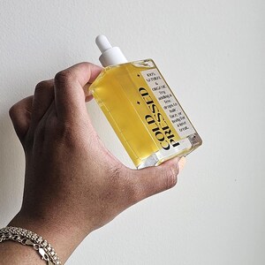May include: A clear glass bottle with a white dropper top filled with a yellow liquid. The bottle is labeled "COLD PRESSED" and "100% NATURAL & ORGANIC". The label also includes instructions for using the oil, suggesting adding a few drops to hair, face, or body for a "luxe treat."