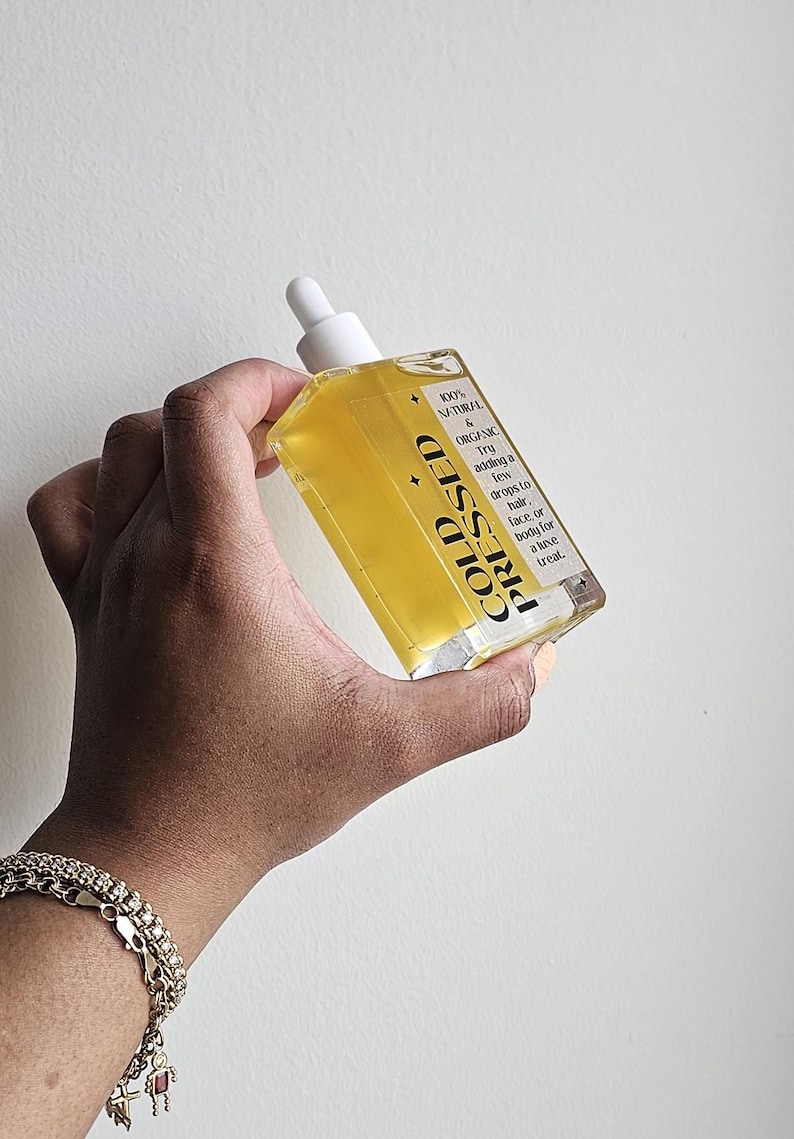 May include: A clear glass bottle with a white dropper top filled with a yellow liquid. The bottle is labeled "COLD PRESSED" and "100% NATURAL & ORGANIC". The label also includes instructions for using the oil, suggesting adding a few drops to hair, face, or body for a "luxe treat."