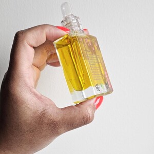 Puede incluir: Una botella de vidrio transparente con un l&iacute;quido amarillo en su interior, sostenida en una mano con esmalte de u&ntilde;as rojo. La botella tiene un cuentagotas transparente y una etiqueta con texto. El fondo es una pared blanca lisa.