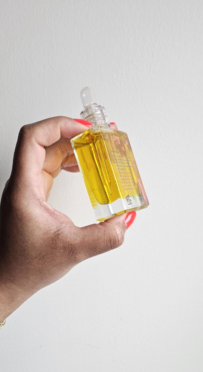 Puede incluir: Una botella de vidrio transparente con un l&iacute;quido amarillo en su interior, sostenida en una mano con esmalte de u&ntilde;as rojo. La botella tiene un cuentagotas transparente y una etiqueta con texto. El fondo es una pared blanca lisa.