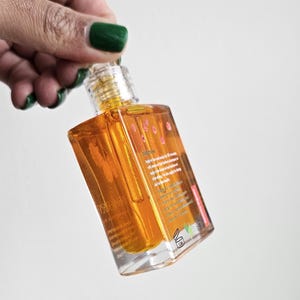 Puede incluir: Una botella transparente de forma cuadrada llena de l&iacute;quido dorado. La botella tiene una tapa transparente y una etiqueta con texto. La botella est&aacute; sostenida por una mano con esmalte de u&ntilde;as verde. El fondo es blanco liso.