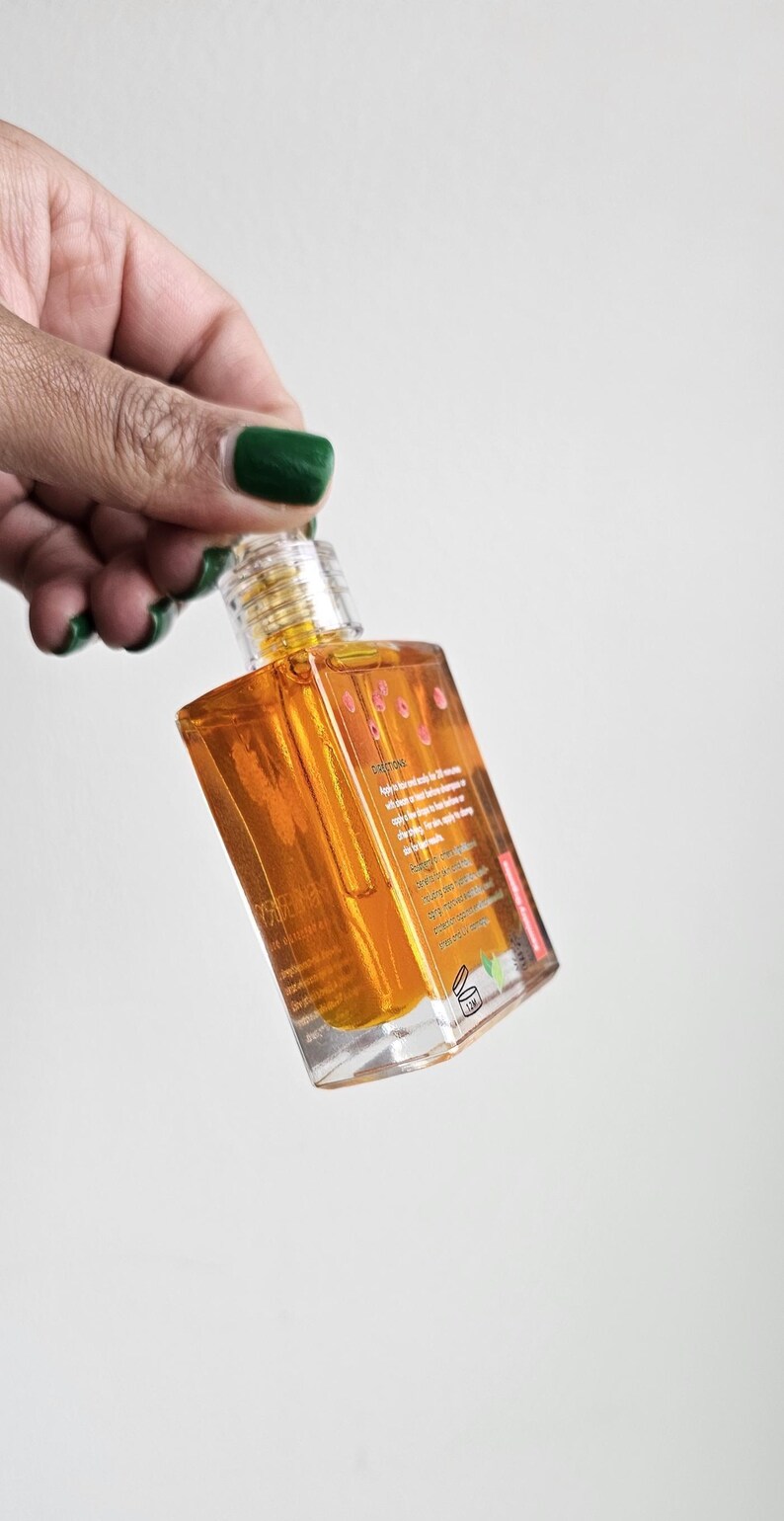 Puede incluir: Una botella transparente de forma cuadrada llena de l&iacute;quido dorado. La botella tiene una tapa transparente y una etiqueta con texto. La botella est&aacute; sostenida por una mano con esmalte de u&ntilde;as verde. El fondo es blanco liso.