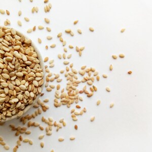 May include: A close-up shot of a small, light-colored bowl filled with sesame seeds. Some seeds are scattered on the white surface around the bowl. The seeds are small, oval-shaped, and have a light tan color.