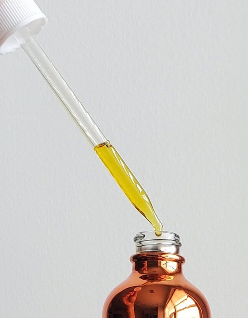 Pode incluir: Uma garrafa de vidro cor de cobre com gargalo prateado e um conta-gotas de vidro transparente. O conta-gotas est&aacute; cheio de um l&iacute;quido amarelo, com uma gota a formar-se na ponta. A garrafa est&aacute; sobre um fundo branco.