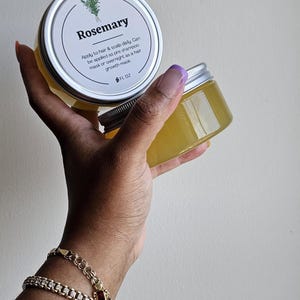 May include: Two clear jars of Rosemary hair product, each with a silver lid and a white label. The label includes the word "Rosemary" and instructions for use. The jars are held in a hand, with a gold bracelet visible.