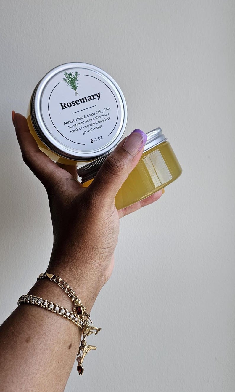 May include: Two clear jars of Rosemary hair product, each with a silver lid and a white label. The label includes the word "Rosemary" and instructions for use. The jars are held in a hand, with a gold bracelet visible.