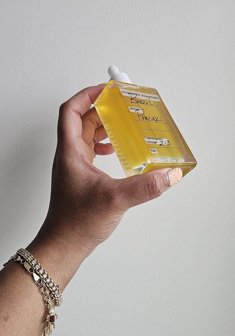 May include: A clear glass bottle with a dropper filled with a yellow liquid. The bottle has a white label with the text "Ayurvedic Essentials Brazil Pracaxi Blend Comedogenic rating: 2-3 Exp"