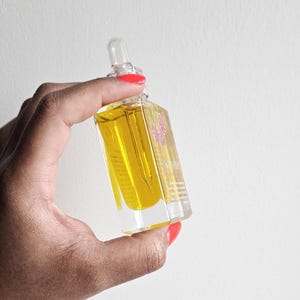 Puede incluir: Una botella de vidrio rectangular transparente llena de l&iacute;quido amarillo, sostenida sobre un fondo blanco. La botella tiene un cuentagotas y una etiqueta floral rosa. La botella es sostenida por una mano con esmalte de u&ntilde;as rojo.
