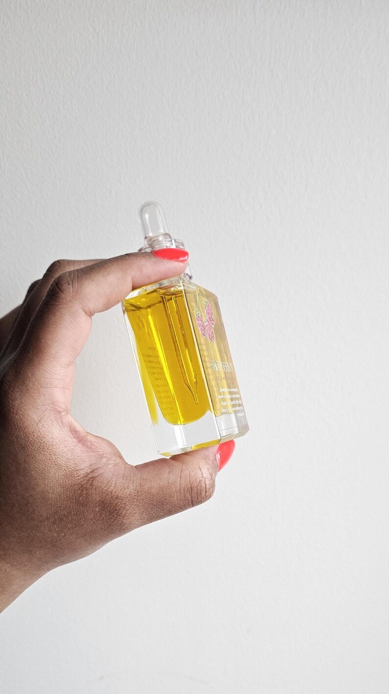 Puede incluir: Una botella de vidrio rectangular transparente llena de l&iacute;quido amarillo, sostenida sobre un fondo blanco. La botella tiene un cuentagotas y una etiqueta floral rosa. La botella es sostenida por una mano con esmalte de u&ntilde;as rojo.