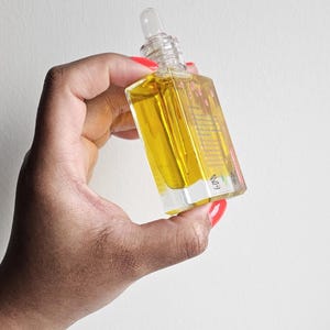 Puede incluir: Una botella de vidrio transparente con un cuentagotas lleno de l&iacute;quido amarillo. La botella tiene forma cuadrada y una tapa transparente. La botella se sostiene en una mano con esmalte de u&ntilde;as rojo y una pulsera dorada. El fondo es una pared blanca lisa.