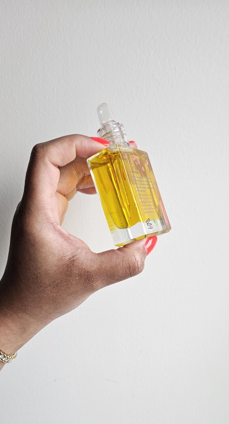 Puede incluir: Una botella de vidrio transparente con un cuentagotas lleno de l&iacute;quido amarillo. La botella tiene forma cuadrada y una tapa transparente. La botella se sostiene en una mano con esmalte de u&ntilde;as rojo y una pulsera dorada. El fondo es una pared blanca lisa.