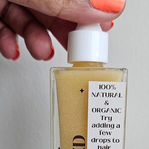 May include: A clear glass bottle with a white dropper top filled with a golden liquid. The label reads "COLD PRESSED" and "100% NATURAL & ORGANIC. Try adding a few drops to hair, face, or body for a luxe treat."