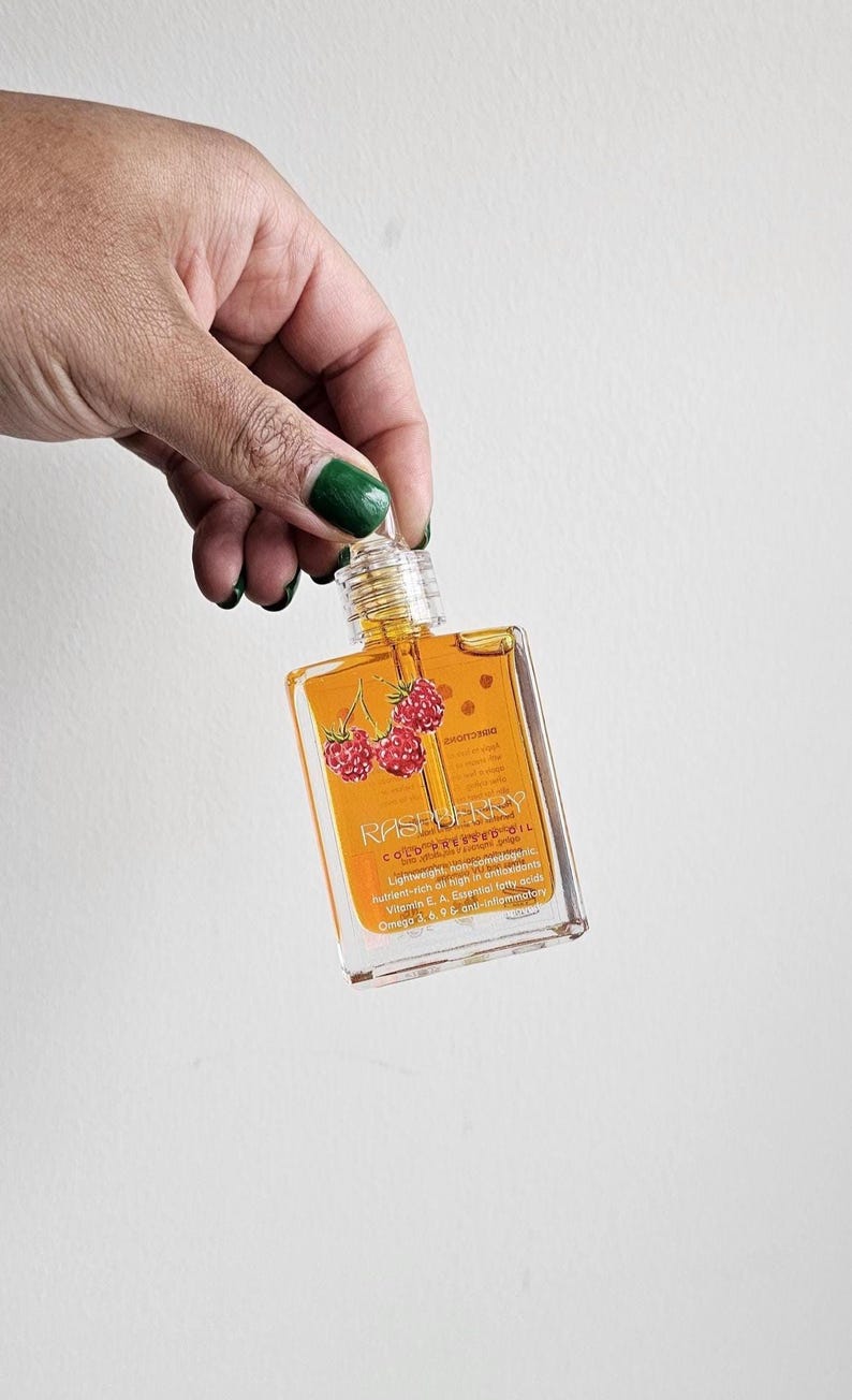 Puede incluir: Una botella rectangular transparente de aceite de frambuesa prensado en fr&iacute;o. La etiqueta presenta la palabra "RASPBERRY" y un gr&aacute;fico de tres frambuesas rojas. La botella est&aacute; sostenida por una mano con esmalte de u&ntilde;as verde.